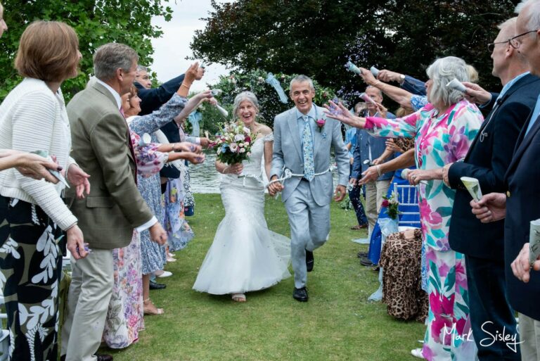 The joyous newlyweds exit the wedding ceremony skipping between their guests to a sea of confetti after their Bisham Abbey outdoor marriage