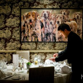 Photography of a member of staff setting up a table in the dining area whilst stood under a huge painting at the Bay Tree Hotel in Burford 