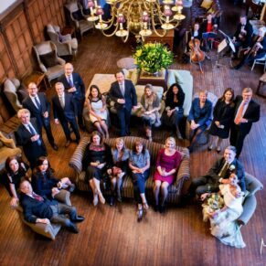 All of the wedding guests gathered in the Great Hall at Danesfield House look towards the balcony above for this group pose