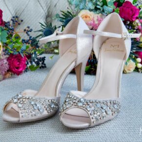 The bride's glimmering shoes placed onto a pale grey chair with her vibrant bouquet 
