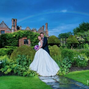 The view through the Chenies Manor gardens on a summer's day with the newlyweds framed by a stunning rainbow against darkened skies