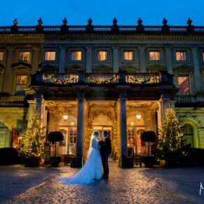 Photography of the newlyweds striking a pose at the front of the floodlit property under a dusky blue sky for their Cliveden House Christmas wedding  