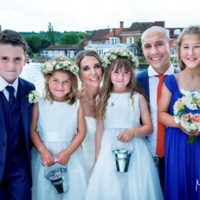 The bride and groom snuggle up to their pageboy and flower girls with the river Thames and their reception venue seen beyond completing this stunning capture