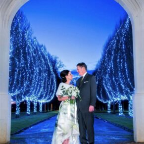 Night shot of the newlyweds under an archway with tree illuminations behind them at their Danesfield House winter wedding  