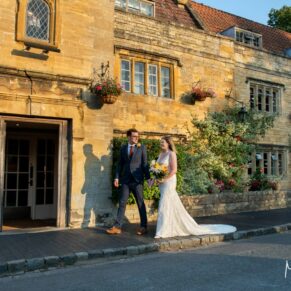 The newlyweds walk past the grand street entrance at their Manor House Hotel wedding reception in Moreton-in-Marsh